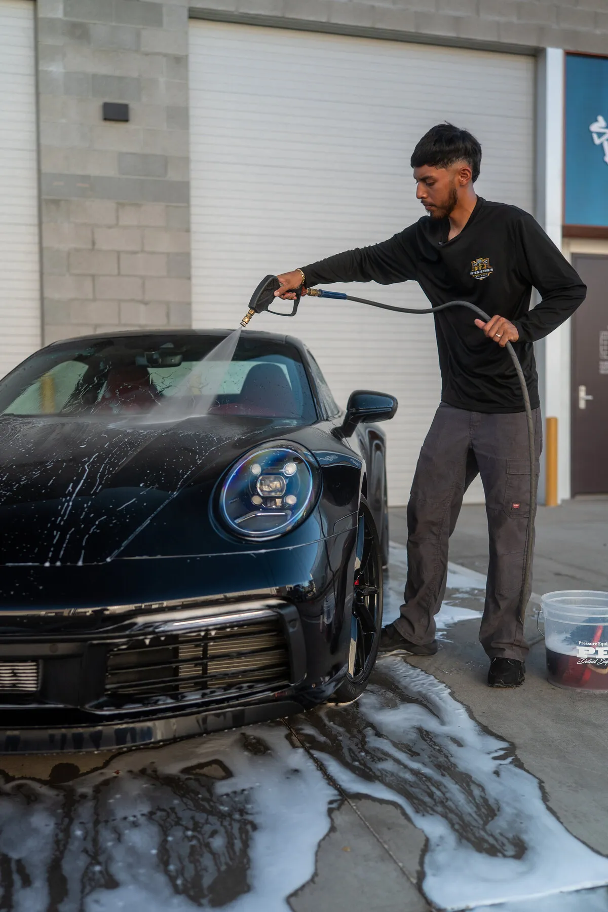 Reyes Details technician rinsing a black Porsche 911 with a pressure washer, foam running off the paint