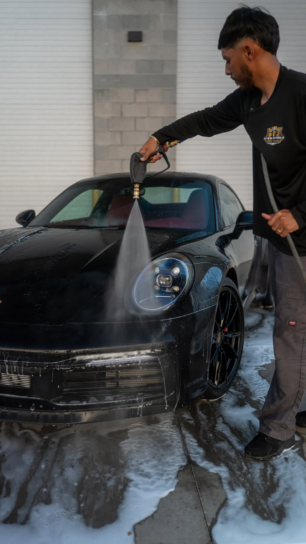 Reyes Details technician spraying down the front end of a black Porsche 911 in an outdoor bay