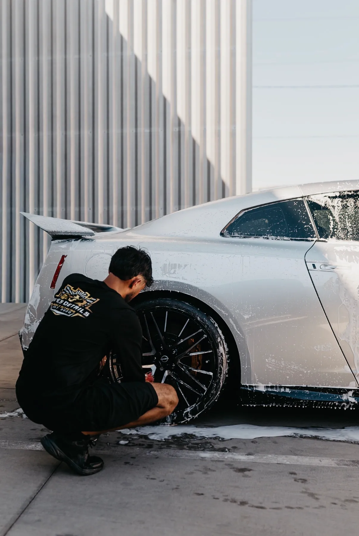 Reyes Details technician crouched at the rear wheel of a foam-covered silver GT-R, cleaning the barrel by hand