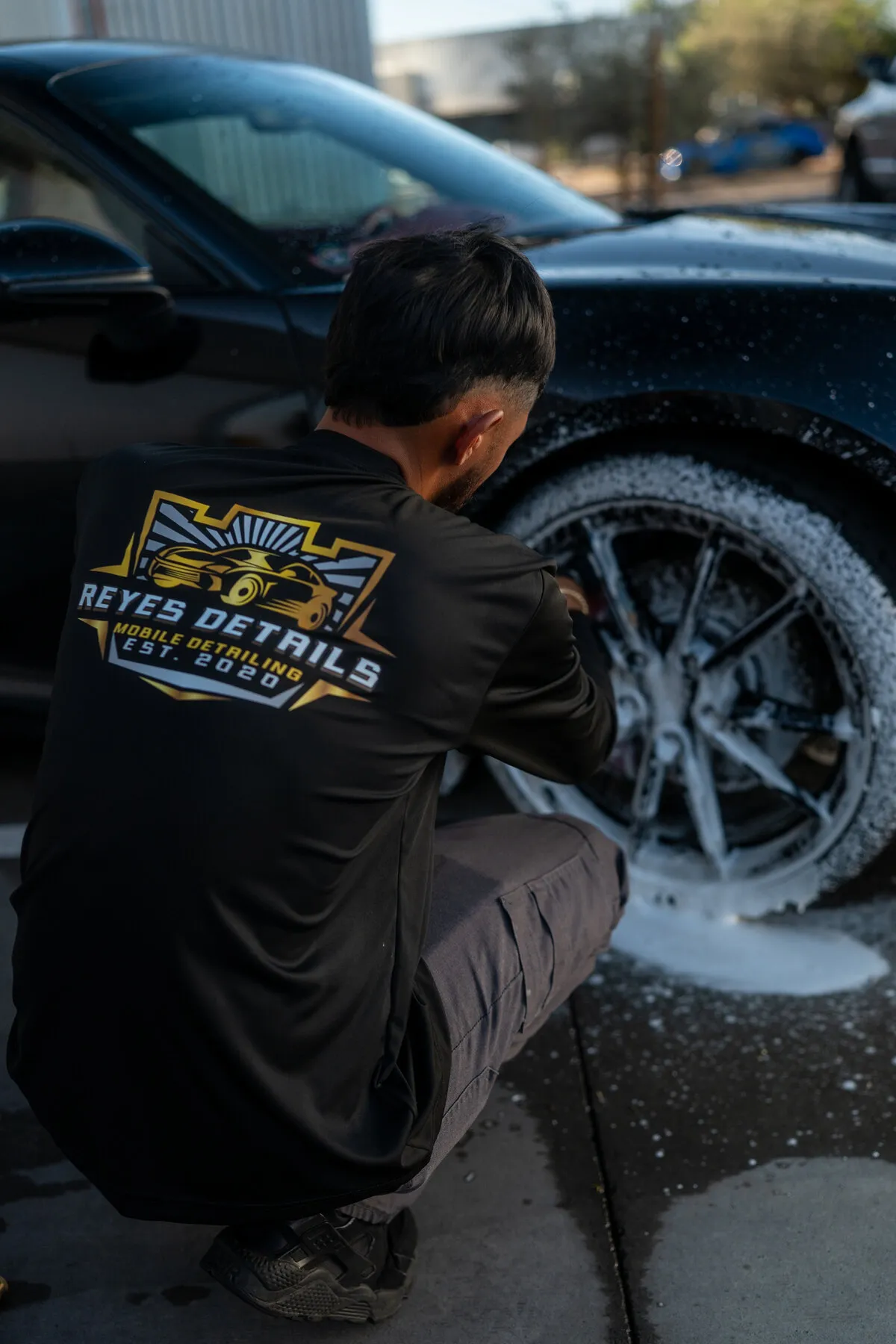 Close view of a Reyes Details technician in a branded shirt scrubbing the wheel of a black Porsche 911